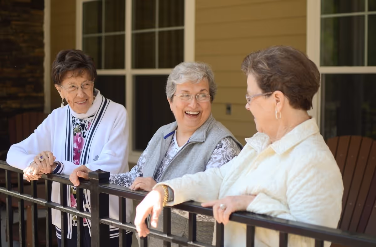 Three elderly women standing outside on a porch or patio, leaning on a black metal railing and engaging in a cheerful conversation. They are dressed in light jackets and sweaters, with a building featuring large windows in the background.