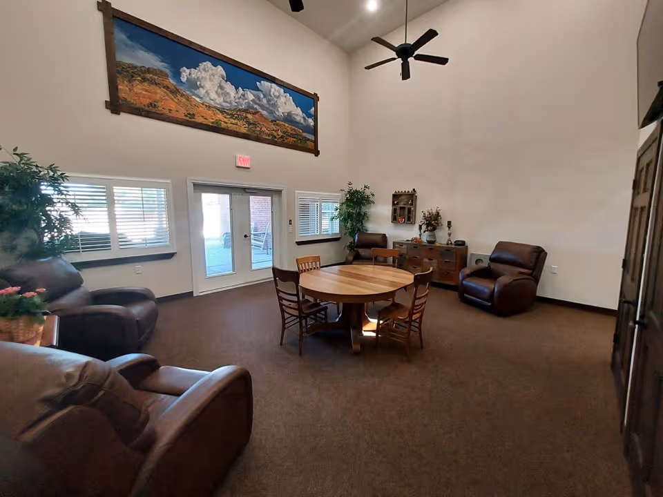 A spacious living room with high ceilings featuring a ceiling fan, four brown leather armchairs, a round wooden table with four chairs, two windows with blinds, a glass door leading outside, a wooden cabinet with decorative items, and a large landscape painting on the wall above the door.