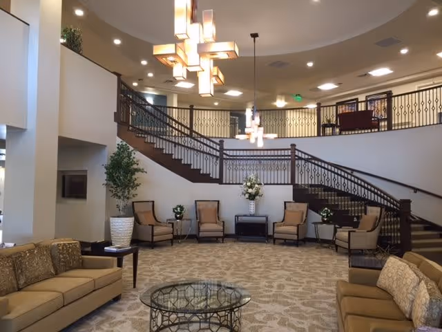 Spacious and elegant interior common area with beige sofas, armchairs, a glass coffee table, decorative plants, and a large staircase with ornate railing leading to an upper level. Modern pendant lights hang from the ceiling, illuminating the space.