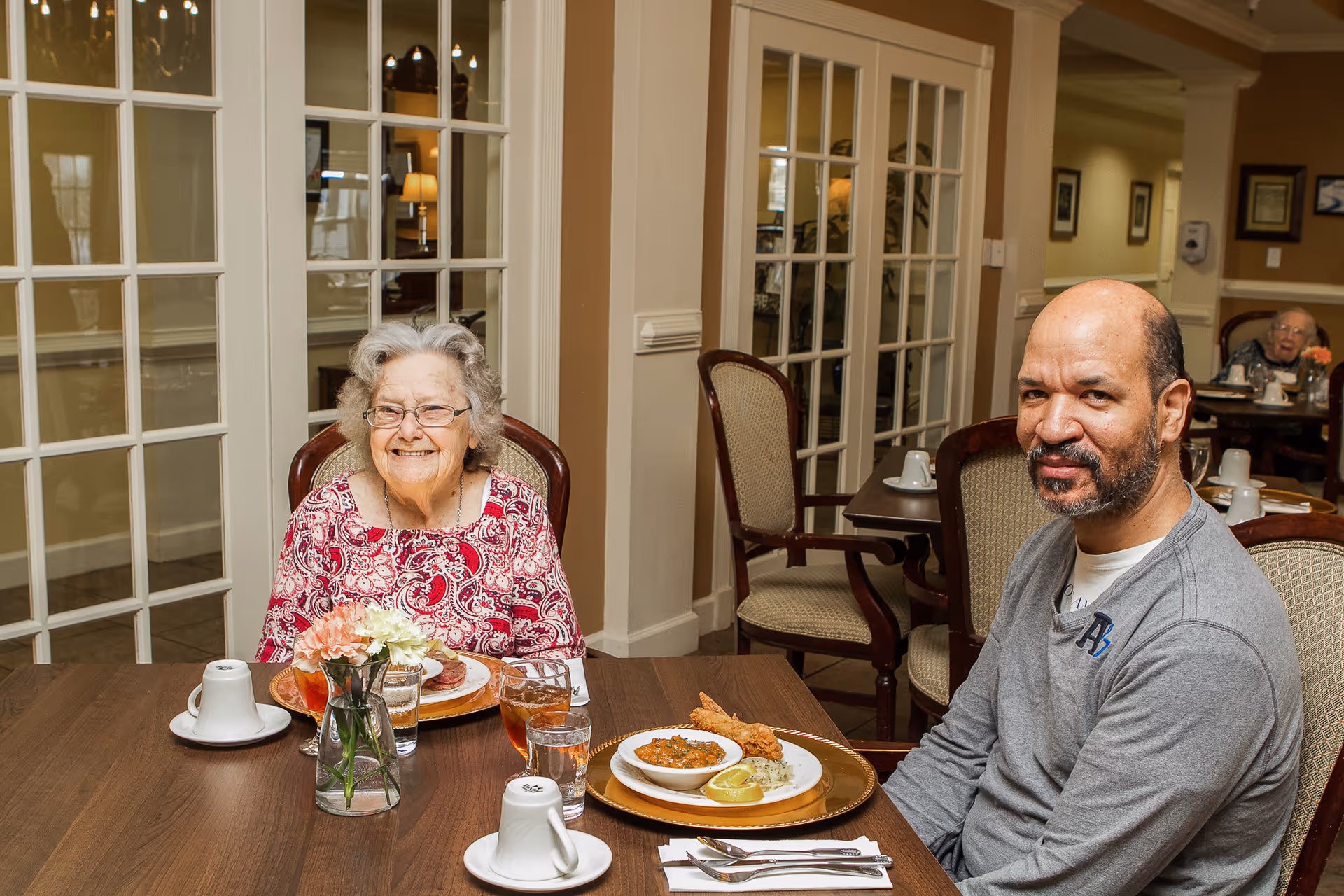 An elderly woman and a middle-aged man sitting at a dining table in a well-lit room with glass-paneled doors in the background. The table is set with plates of food, glasses of iced tea, and a small vase with flowers. Both individuals are smiling and appear to be enjoying their meal.