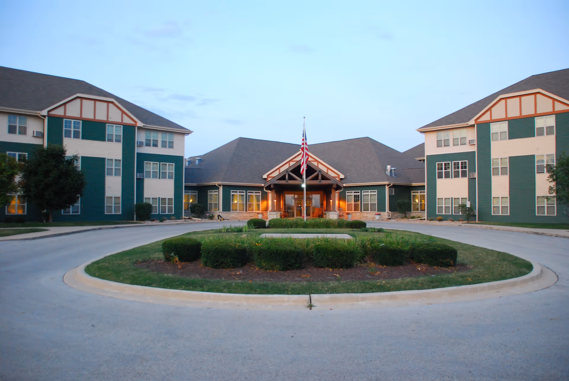 Exterior view of The Timbers of Shorewood senior living facility at dusk, showing a circular driveway with landscaped bushes and an American flag in the center, flanked by two three-story buildings with green and beige siding and multiple windows.