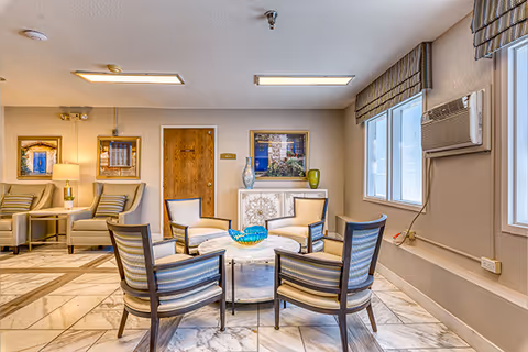 A cozy senior living facility common area with a circular arrangement of five cushioned chairs around a round marble table holding a decorative blue glass bowl. The room has beige walls, two framed pictures, a wooden door, and a window with an air conditioning unit. Additional seating with armchairs and a lamp is visible in the background.