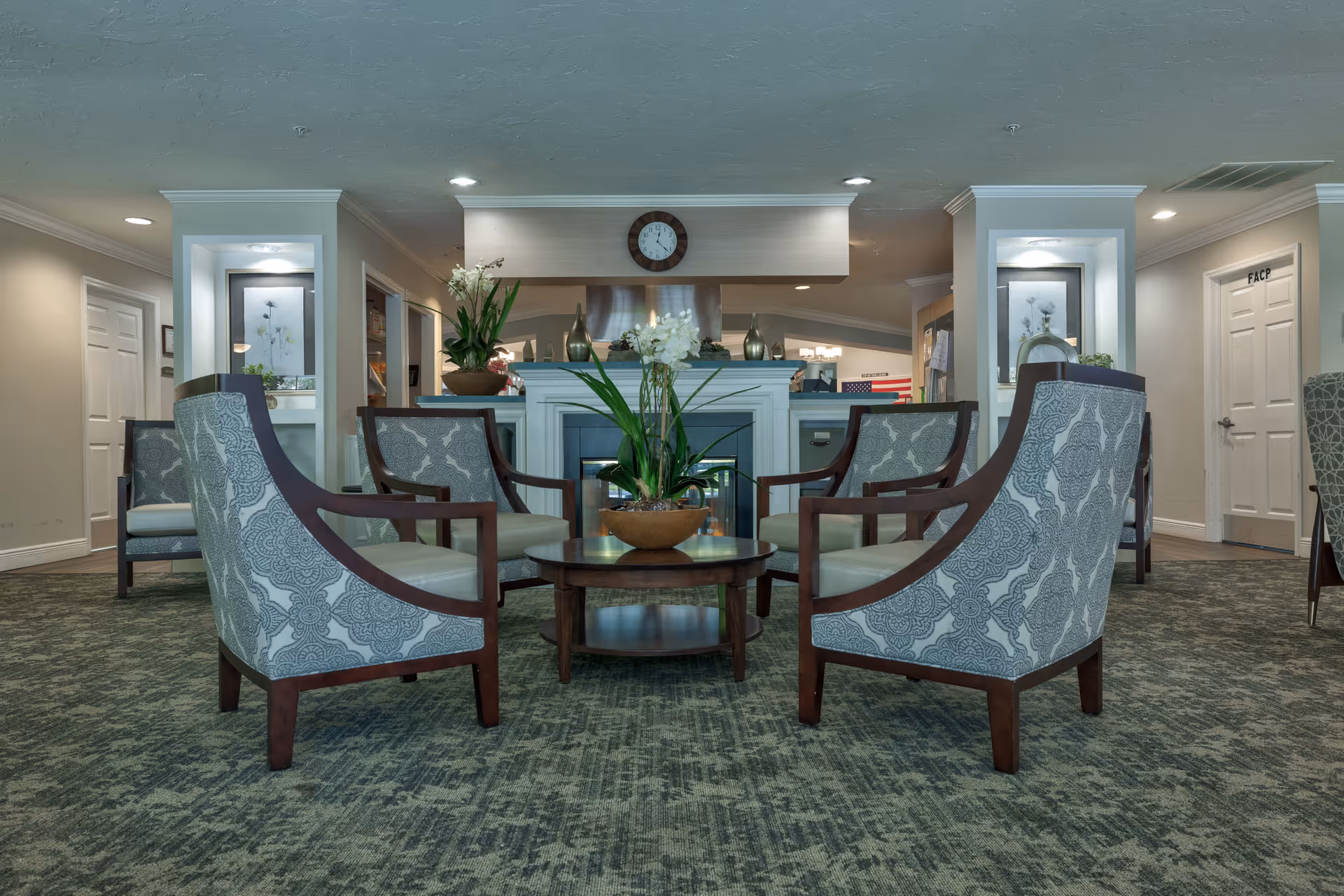 A cozy seating area in a senior living facility with six patterned armchairs arranged around a round wooden coffee table with a potted plant. The room features a fireplace with decorative vases and flowers on the mantel, a clock above it, and framed artwork on the walls. The carpet is green with a textured pattern, and the walls are painted in neutral tones with recessed lighting.