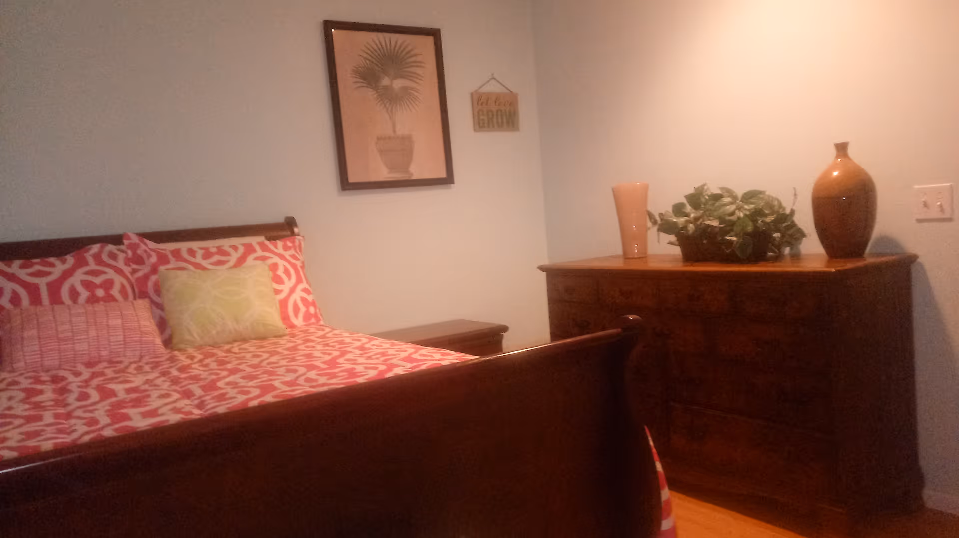 A cozy bedroom with a wooden sleigh bed dressed in pink patterned bedding, a wooden dresser topped with vases and a plant, and framed wall art on a pale blue wall.