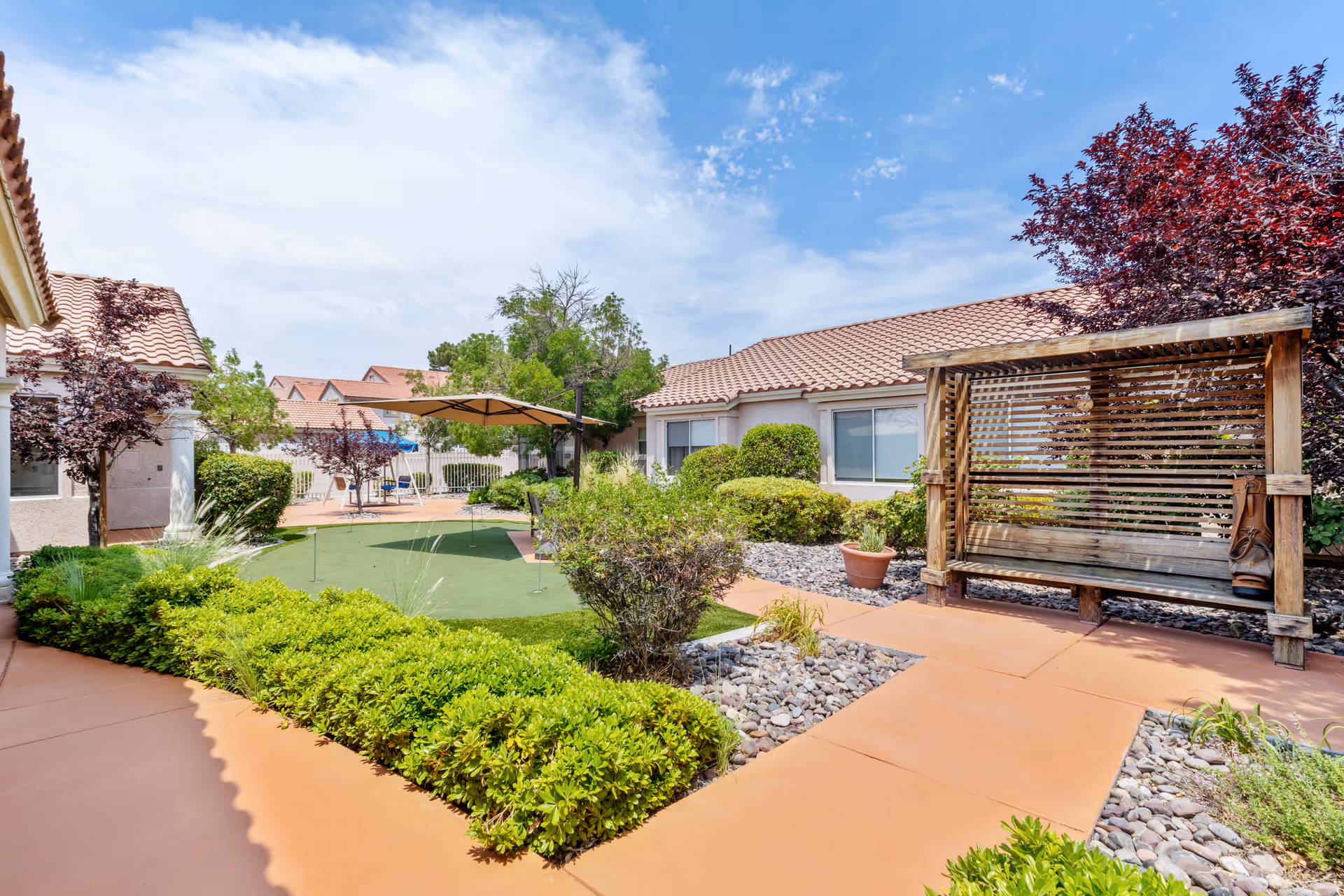 Outdoor courtyard area of a senior living facility with a putting green, shaded seating area with an umbrella, a wooden swing bench, various bushes and trees, and buildings with tiled roofs under a partly cloudy sky.