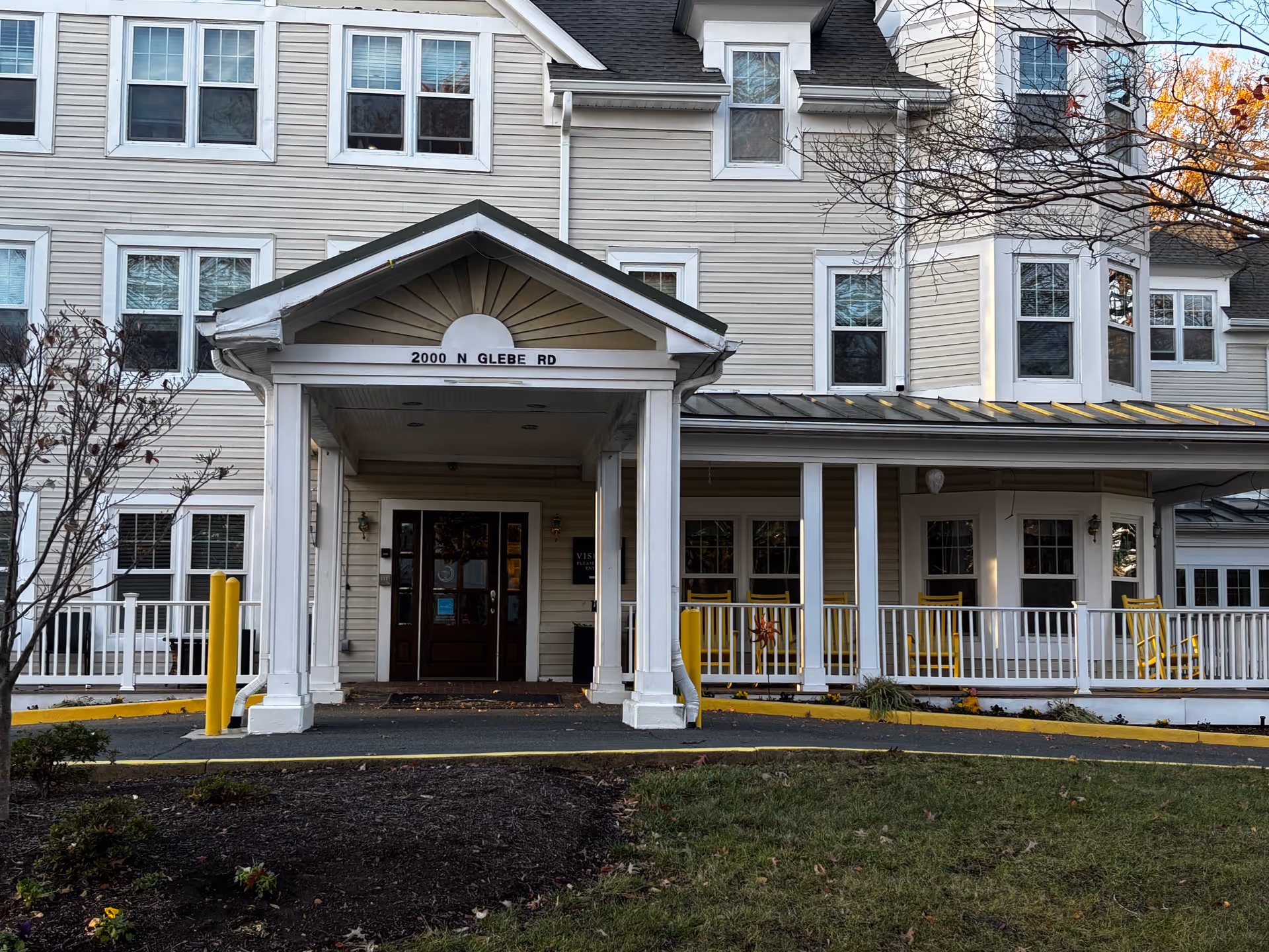 Front entrance of a beige multi-story building with a covered portico reading "2000 N Glebe Rd" and a wraparound porch with yellow chairs.