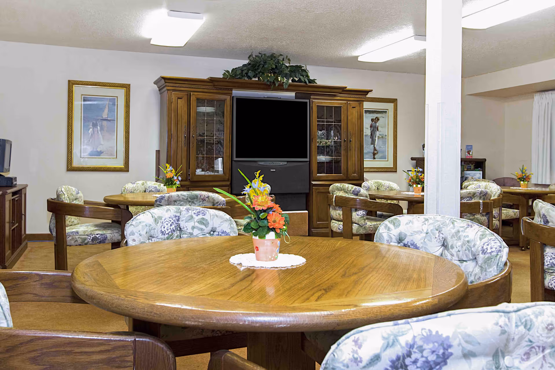 A cozy common area with multiple round wooden tables surrounded by cushioned chairs with floral upholstery. Each table has a small flower arrangement in a pot. In the background, there is a large wooden entertainment center with a television and decorative plants on top. The walls are adorned with framed artwork, and the room is lit by ceiling lights.