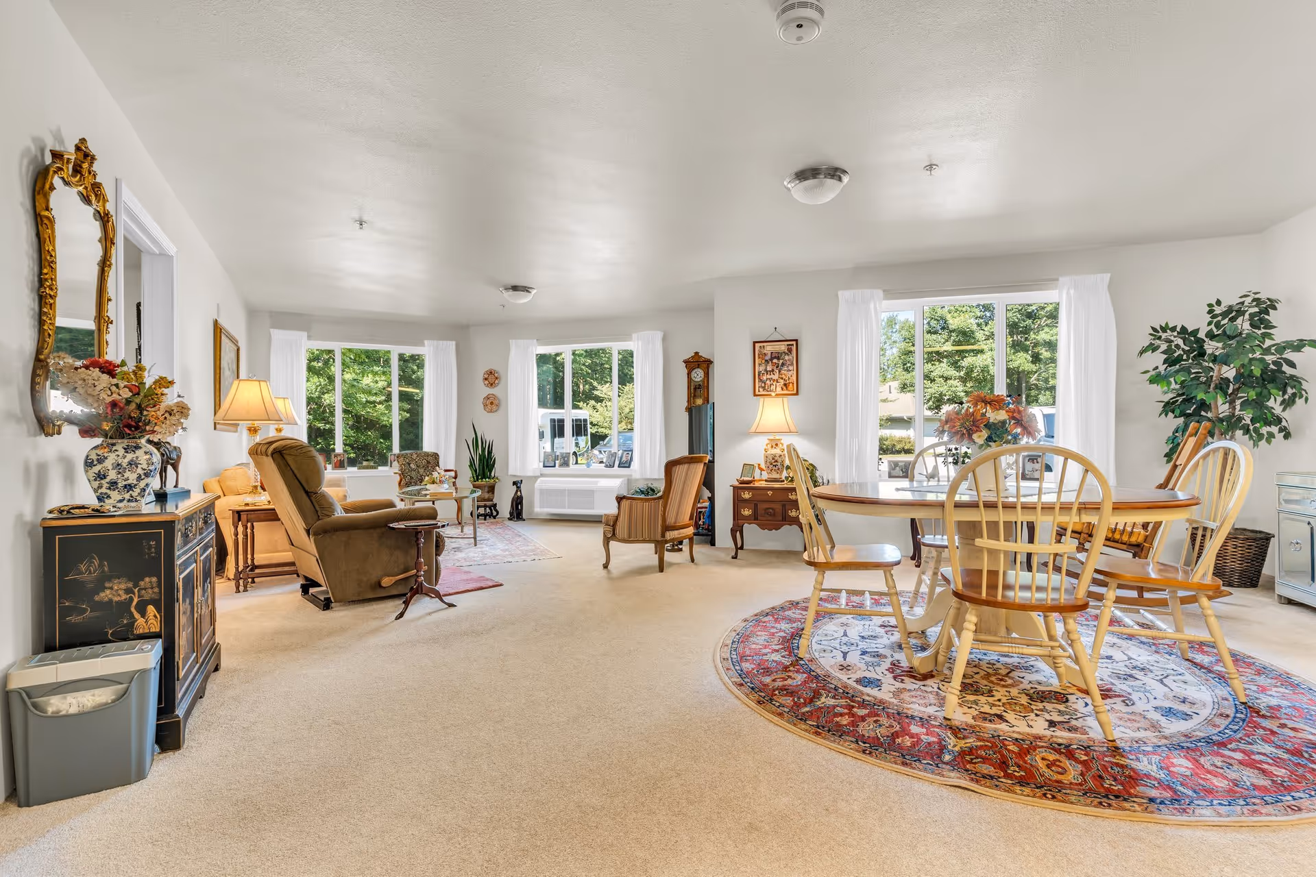 A spacious and well-lit senior living common area featuring a round wooden dining table with six chairs on a colorful patterned rug. The room has large windows with white curtains allowing natural light to fill the space. There are several comfortable armchairs and a sofa arranged around a coffee table near the windows. Decorative elements include a tall grandfather clock, a large potted plant, a side table with a lamp, a wall mirror, and framed artwork. The carpet is beige, and the walls are painted white.