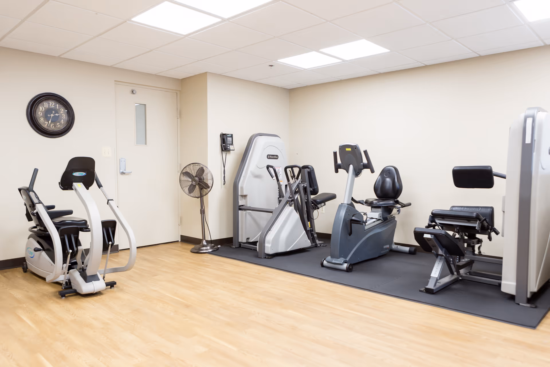 Small fitness room with several exercise machines on a rubber mat, a wall clock, and a standing fan.