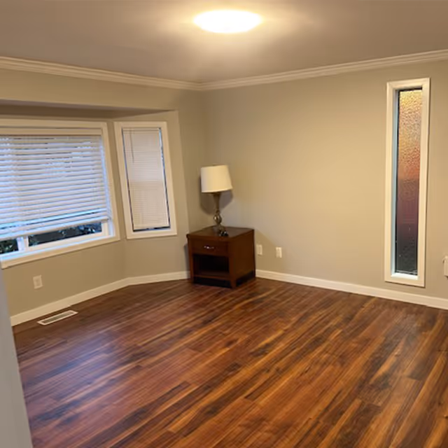 Empty interior room with wood floors, a small side table and lamp beside a bay window and a tall narrow frosted window.