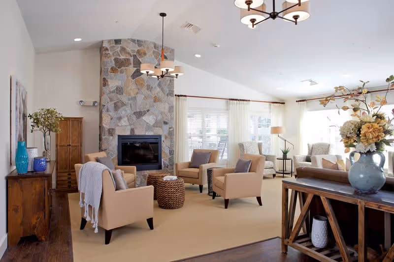 A bright and spacious living room with beige armchairs arranged around a stone fireplace. The room features large windows with white curtains, a wooden sideboard with decorative vases and plants, a wooden console table with a large floral arrangement, and modern ceiling light fixtures.