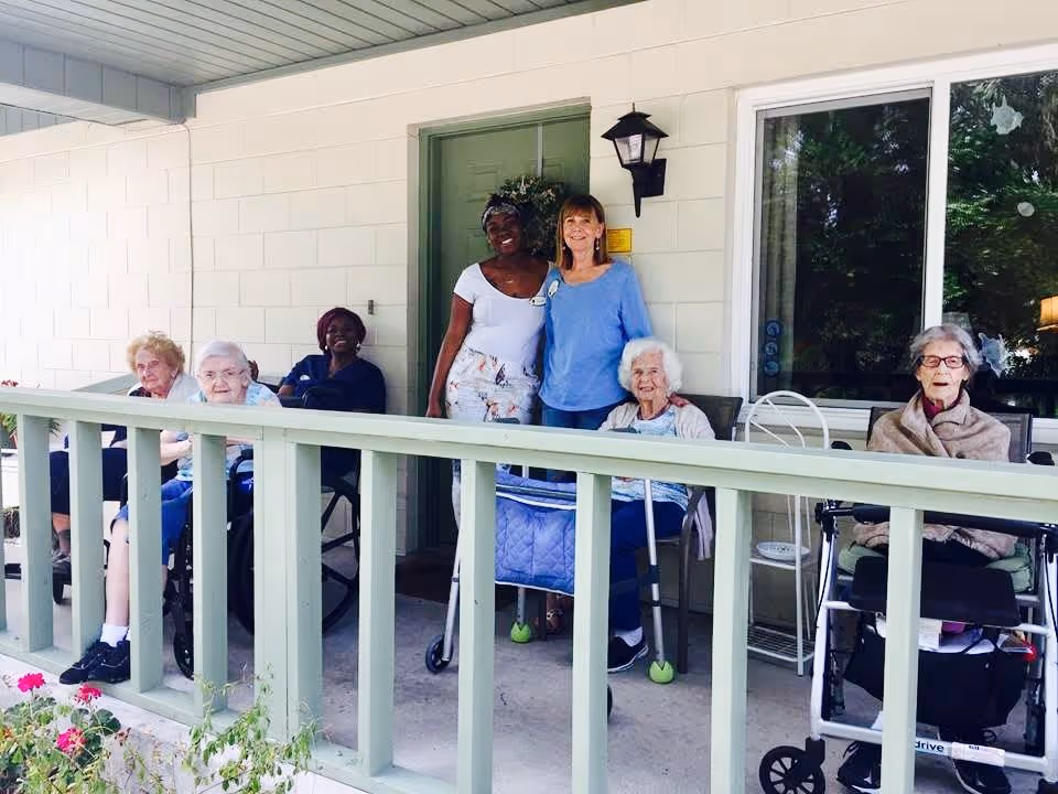 A group of elderly women and two caregivers are gathered on a covered porch outside a building. Some of the elderly women are seated in wheelchairs and using walkers, while the caregivers stand behind them smiling. The porch has a green railing and a green door with a wreath. There is a window reflecting greenery on the right side.