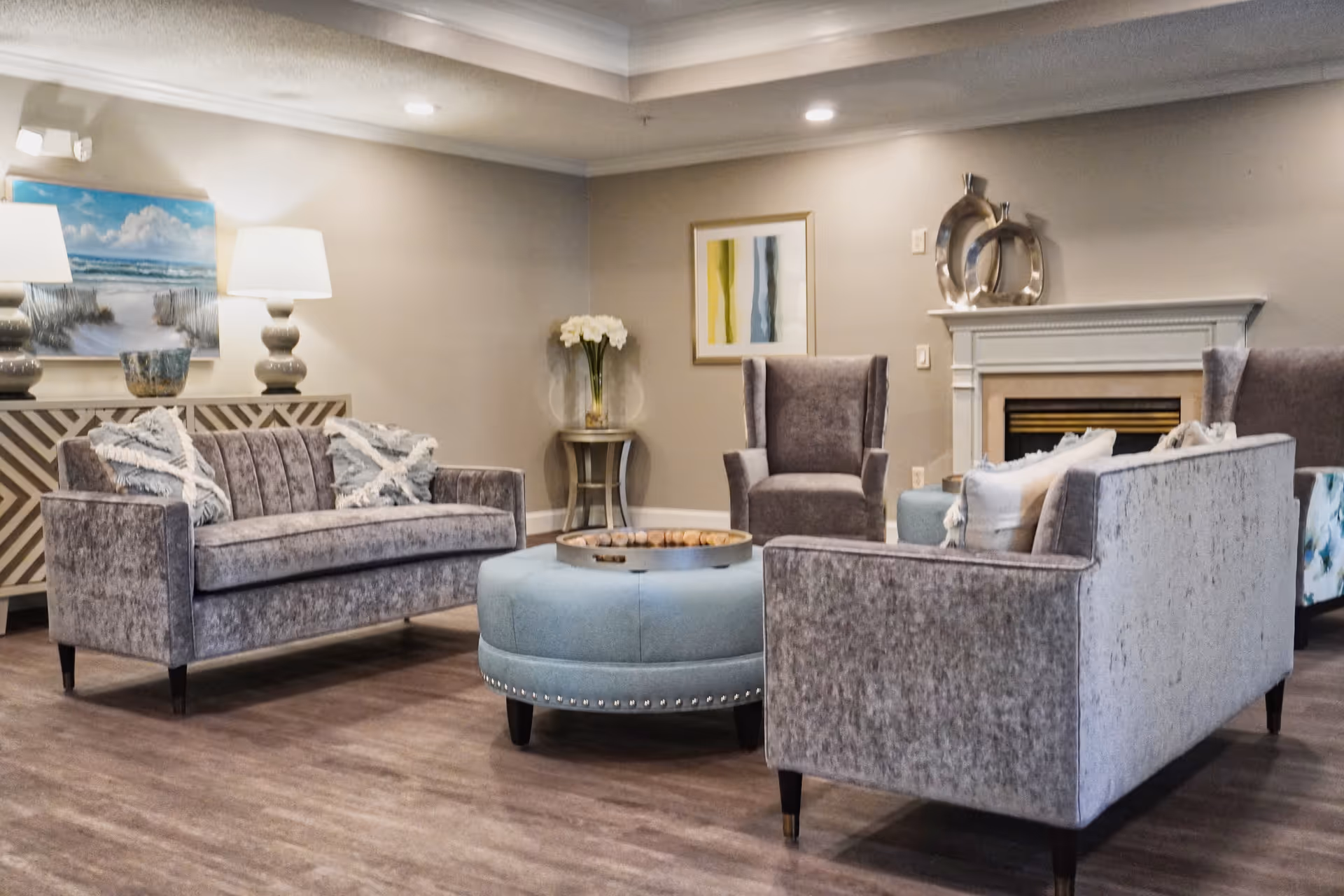 Elegant lounge with gray sofas and armchairs arranged around a round blue ottoman in front of a fireplace.