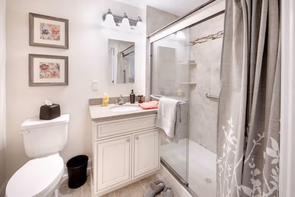 A clean and bright bathroom featuring a white toilet with a black tissue box on top, a white vanity with a sink and countertop holding soap and a towel, a mirror with three light fixtures above it, and a glass-enclosed shower with a gray and white floral shower curtain partially drawn to the side.