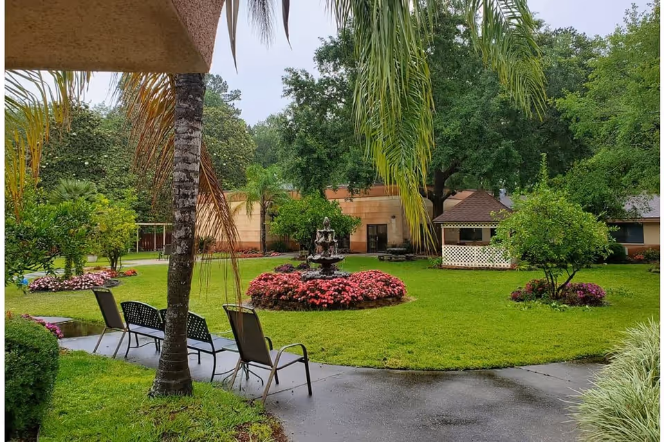 A lush outdoor garden area with green grass, palm trees, and various flowering plants. There is a circular flower bed with pink flowers surrounding a tiered water fountain in the center. Several chairs and a bench are placed along a curved concrete pathway. In the background, there are buildings and a small gazebo-like structure.