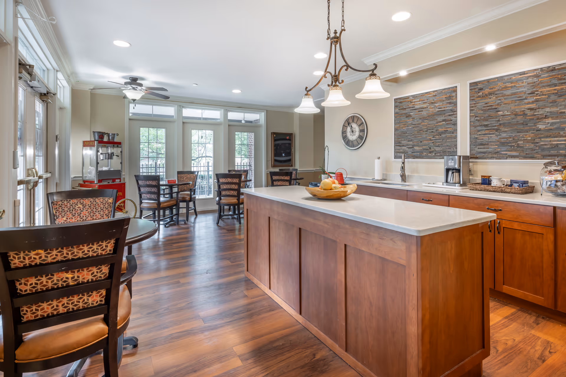 A bright and spacious dining area with wooden flooring, several tables and chairs, a kitchen island with a fruit bowl, a coffee maker, and a popcorn machine near large windows letting in natural light.