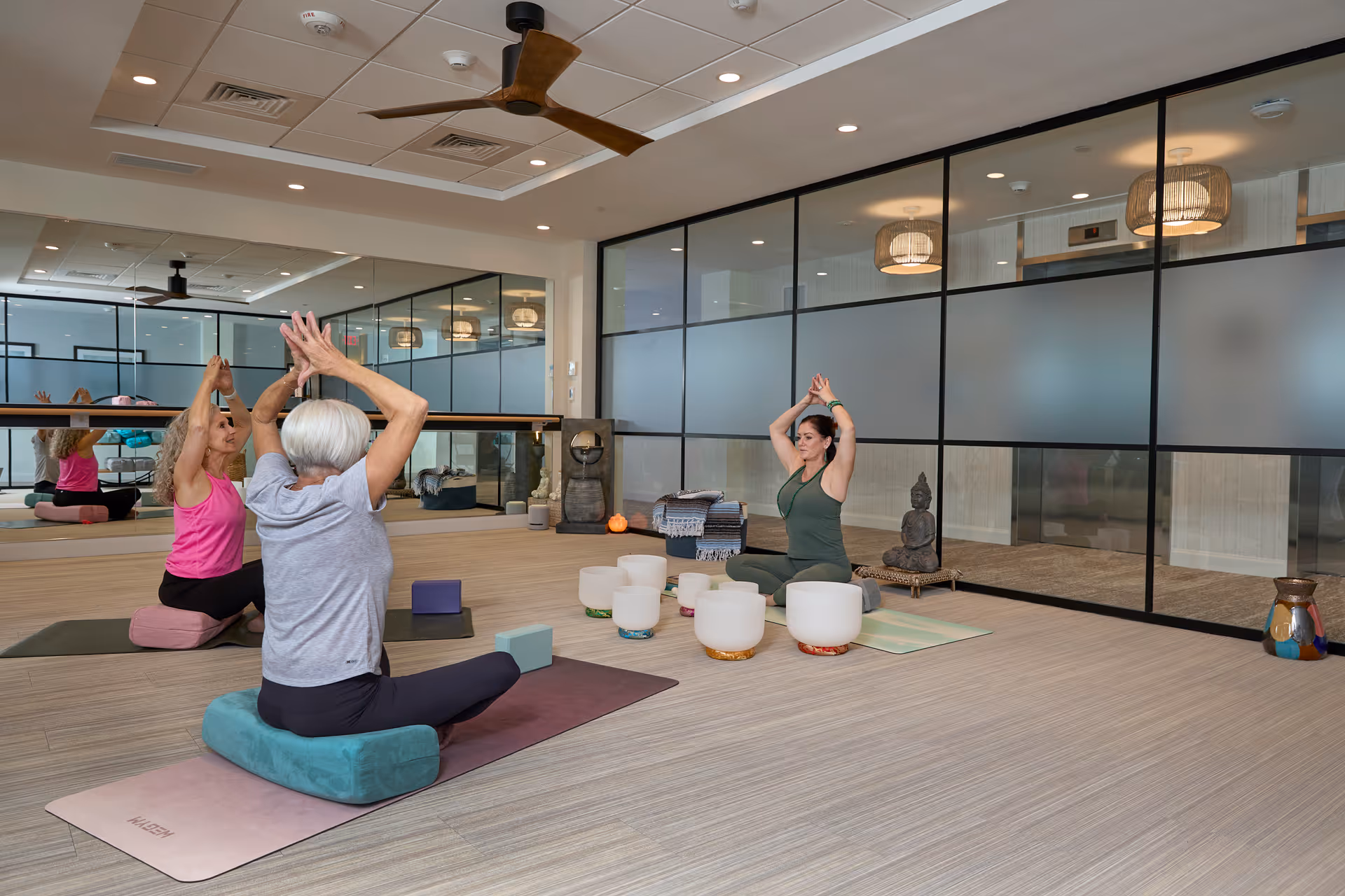Three women practicing yoga in a spacious, well-lit room with wooden floors and large mirrors on one wall. They are seated on yoga mats with cushions, performing a seated stretch with arms raised above their heads. The room has modern decor including a ceiling fan, a Buddha statue, and several white sound bowls arranged on the floor.