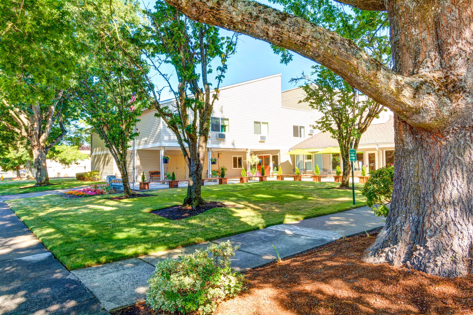 Front exterior of a two-story retirement and assisted living building with a shaded lawn, trees, walkways, and seating areas.