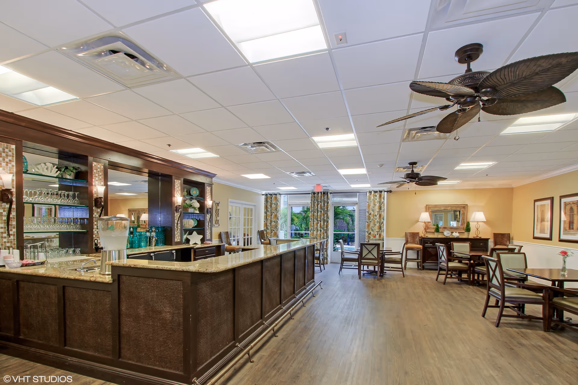 Communal dining and lounge area with a long bar, multiple dining tables, and ceiling fans.