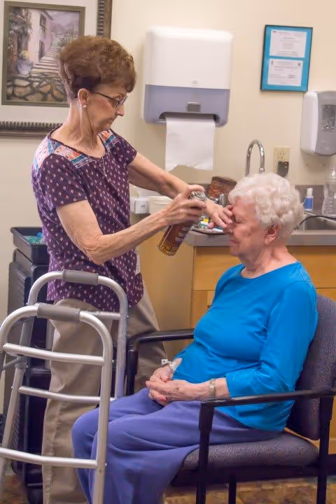 A caregiver sprays a product near the face of an elderly woman seated in a chair inside a room with medical supplies and a walker nearby.