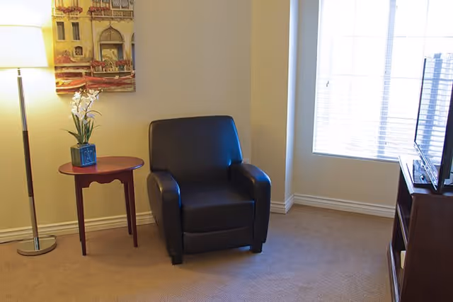 A cozy corner of a living room with a black leather armchair, a small round wooden side table with a potted white orchid, a tall floor lamp, a painting on the wall, and a window with blinds letting in natural light.