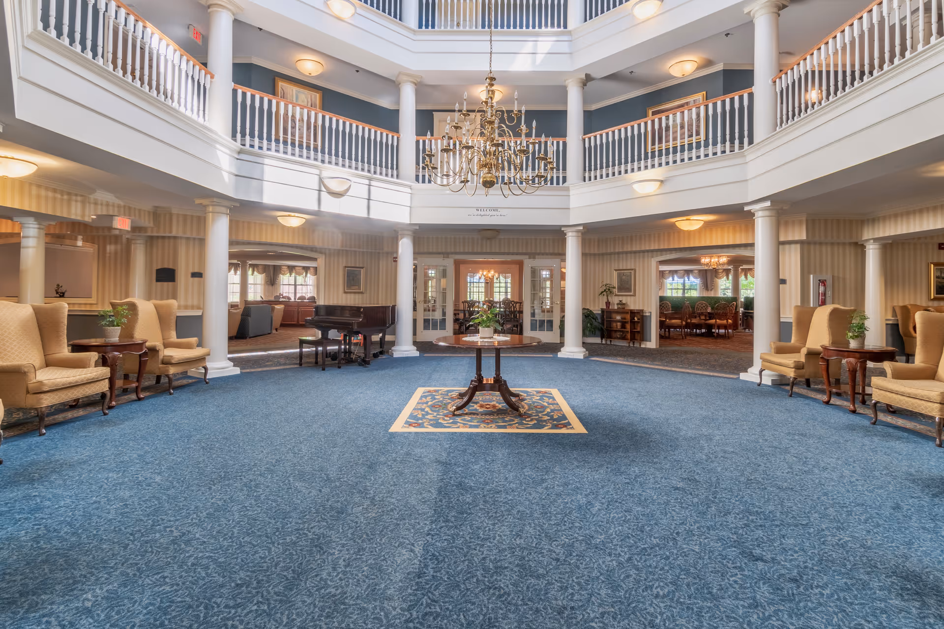Spacious and elegant common area in a senior living facility with blue carpet, beige armchairs arranged along the walls, a central round table with a plant, white columns supporting a second-floor balcony, and a large chandelier hanging from the ceiling.
