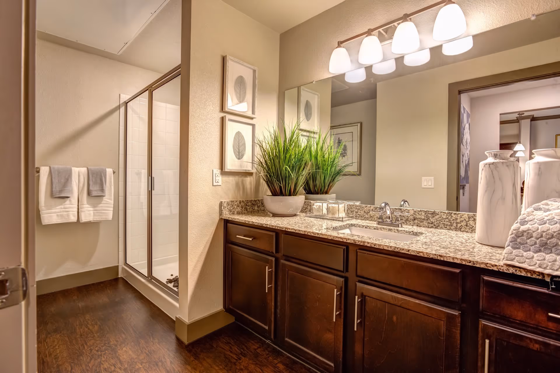 A bathroom with a granite countertop vanity featuring a sink, a large mirror, and decorative plants. There is a glass-enclosed shower in the background and two towels hanging on a rack on the wall. The floor is wooden, and the lighting consists of multiple bulbs above the mirror.