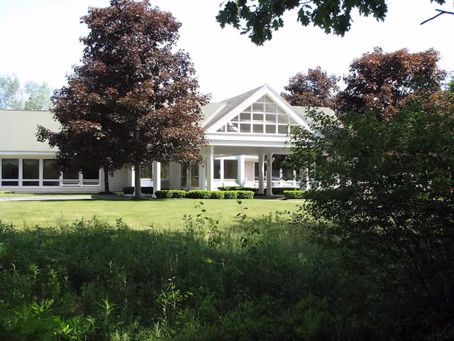 Exterior view of a single-story building with a white facade and large windows, surrounded by green grass, bushes, and trees with reddish-brown leaves under a clear sky.