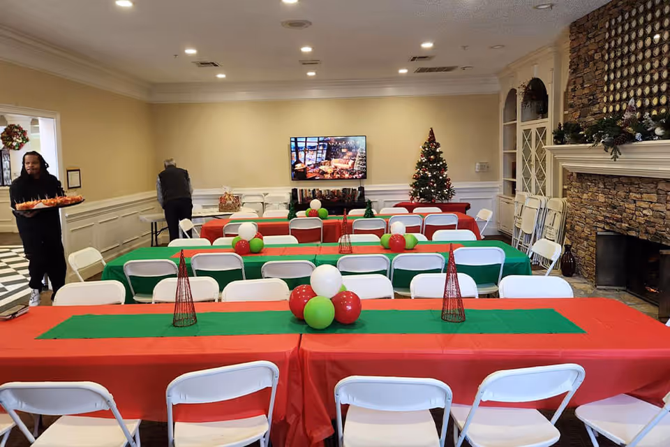 A festive dining room decorated for Christmas with tables covered in red and green tablecloths, white folding chairs, and balloon centerpieces in red, green, and white. A Christmas tree is visible in the back corner next to a TV mounted on the wall. A stone fireplace with holiday decorations is on the right side. Two people are present, one carrying a tray of food and another arranging items on a table.