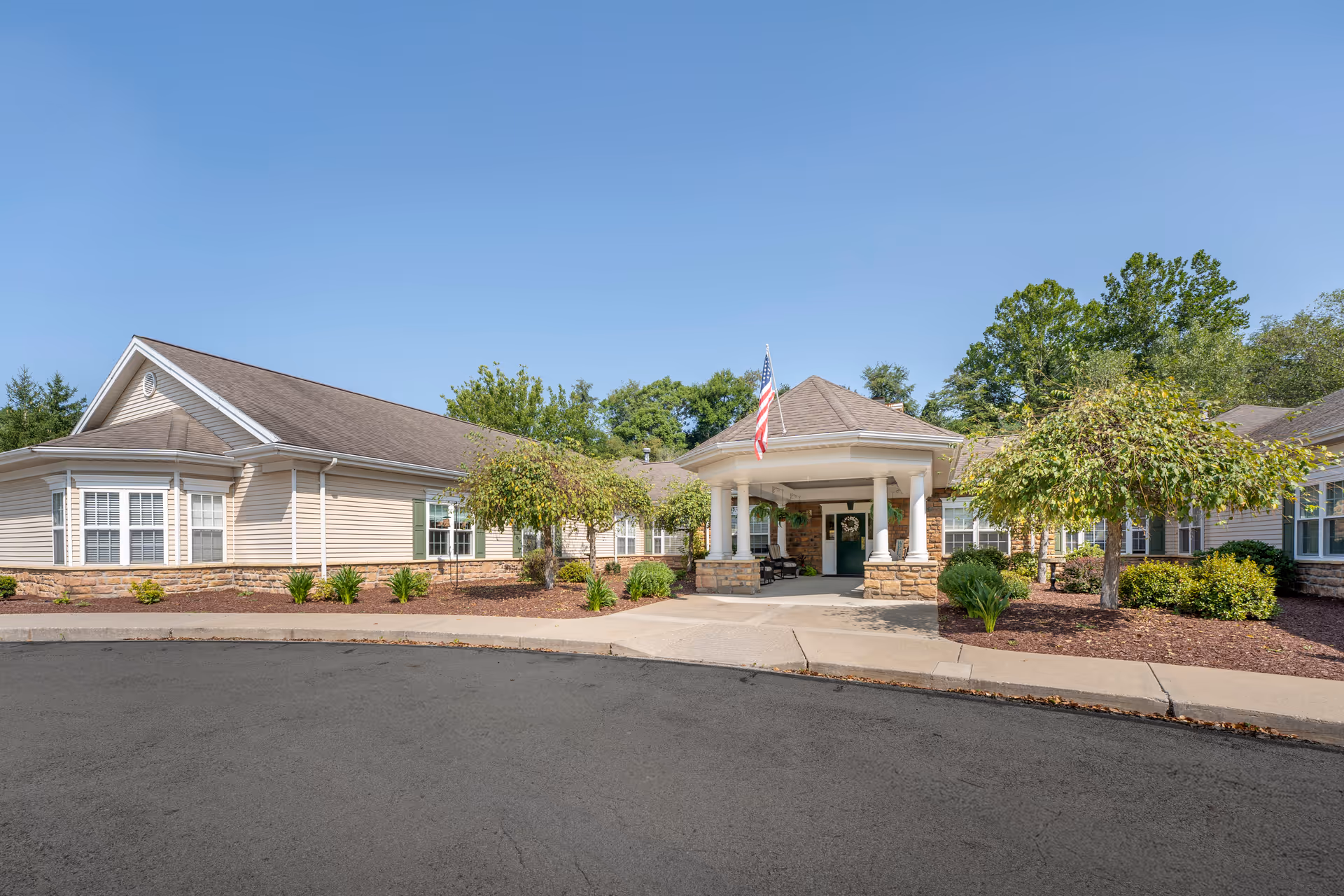Exterior view of a single-story senior living facility building with beige siding and stone accents. The entrance features a covered porch supported by white columns, with an American flag displayed above. There are small trees and landscaped bushes around the building, and a clear blue sky overhead.
