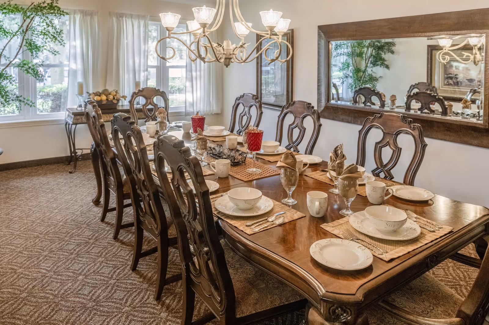 A formal dining room with a long wooden table set for eight people. The table is arranged with plates, bowls, cups, glasses, and napkins. There are ornate wooden chairs around the table, a large mirror on the wall, a chandelier overhead, and windows with white curtains letting in natural light.
