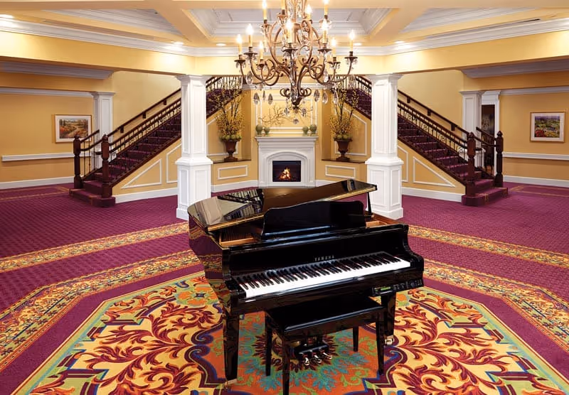 A grand piano sits on a patterned rug in an ornate lobby with twin staircases, chandelier, and a fireplace.