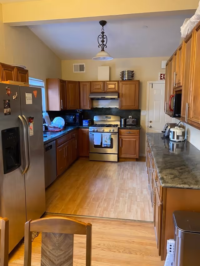 A kitchen with wooden cabinets, stainless steel refrigerator, stove, and dishwasher. The countertops are dark granite, and there are various kitchen appliances such as a microwave, toaster, and rice cooker. The floor is light wood, and a wooden chair is partially visible in the foreground.
