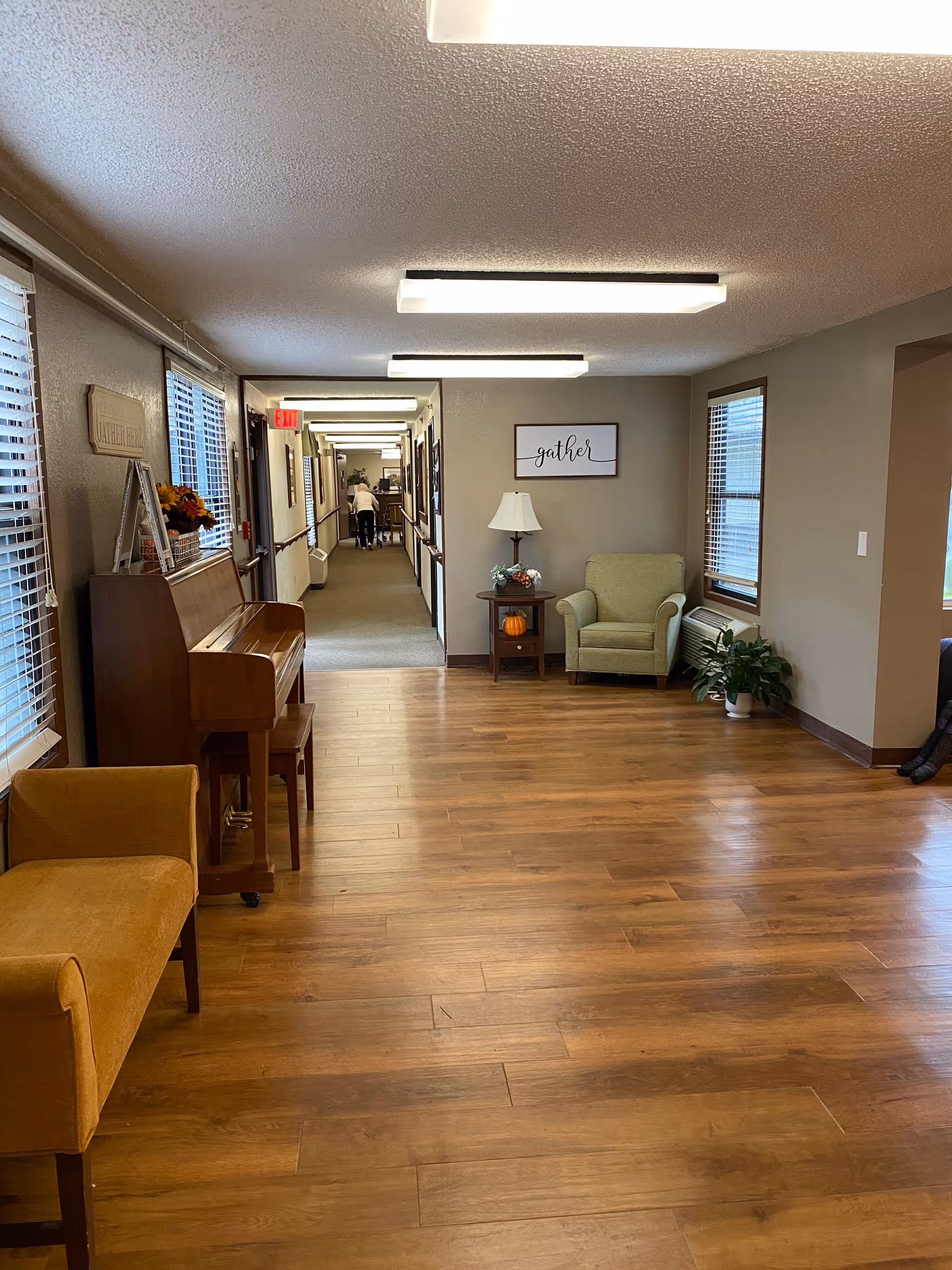 A well-lit hallway in an assisted living facility with wooden flooring and beige walls. On the left side, there is a wooden piano with a small bench and a yellow upholstered bench nearby. On the right side, there is a green armchair next to a small wooden side table with a lamp and decorative items, including a small pumpkin. A framed sign on the wall above the armchair reads 'gather'. The hallway extends into the distance with handrails on both sides and a person walking away pushing a cart. Windows with blinds are visible on both sides.