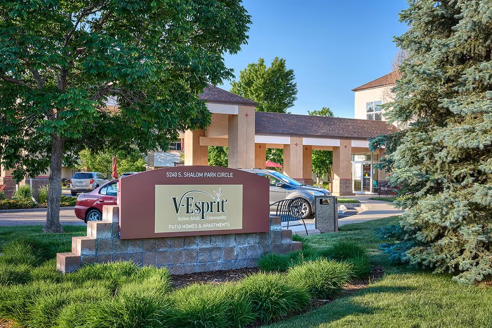 Entrance sign reading "V-Esprit" with landscaped lawn, trees, parked cars, and the facility entrance in the background.