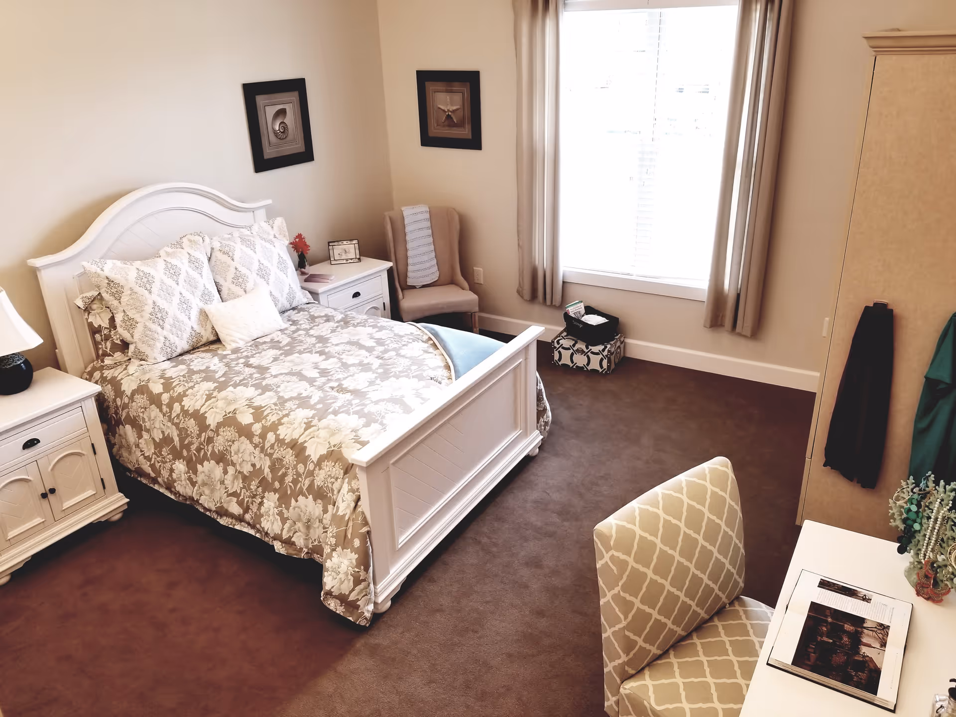A cozy bedroom with a white wooden bed frame and floral patterned bedding. There are two matching nightstands with lamps on either side of the bed. A beige upholstered chair is placed in the corner near a window with beige curtains. A desk with a patterned chair and an open book is positioned near the bottom right corner. The room has beige walls and brown carpet flooring.