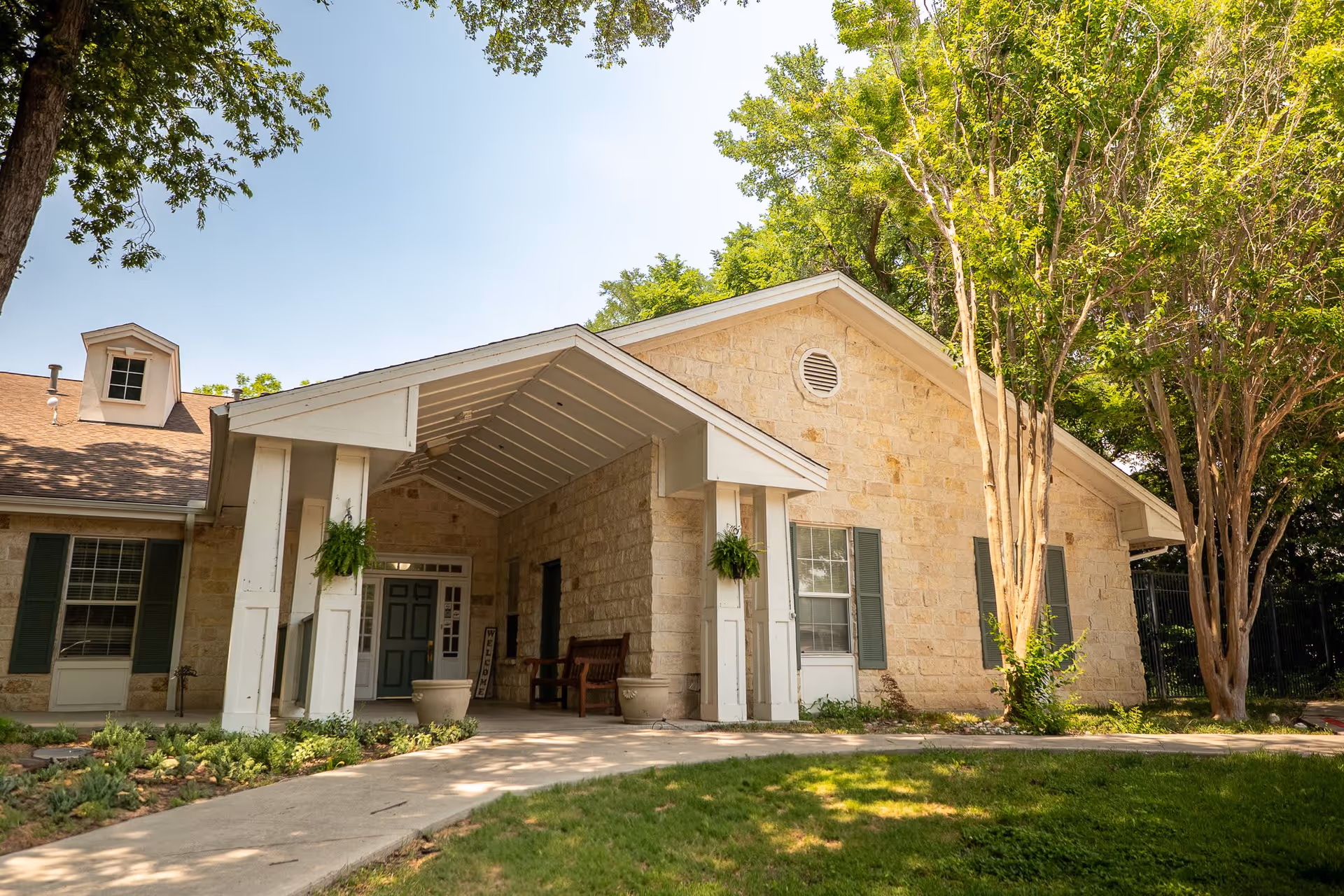 Exterior view of a senior living facility building with light-colored stone walls, green window shutters, and a covered entrance supported by white pillars. There are two hanging plants on the pillars, a wooden bench near the entrance, and trees surrounding the building under a clear blue sky.