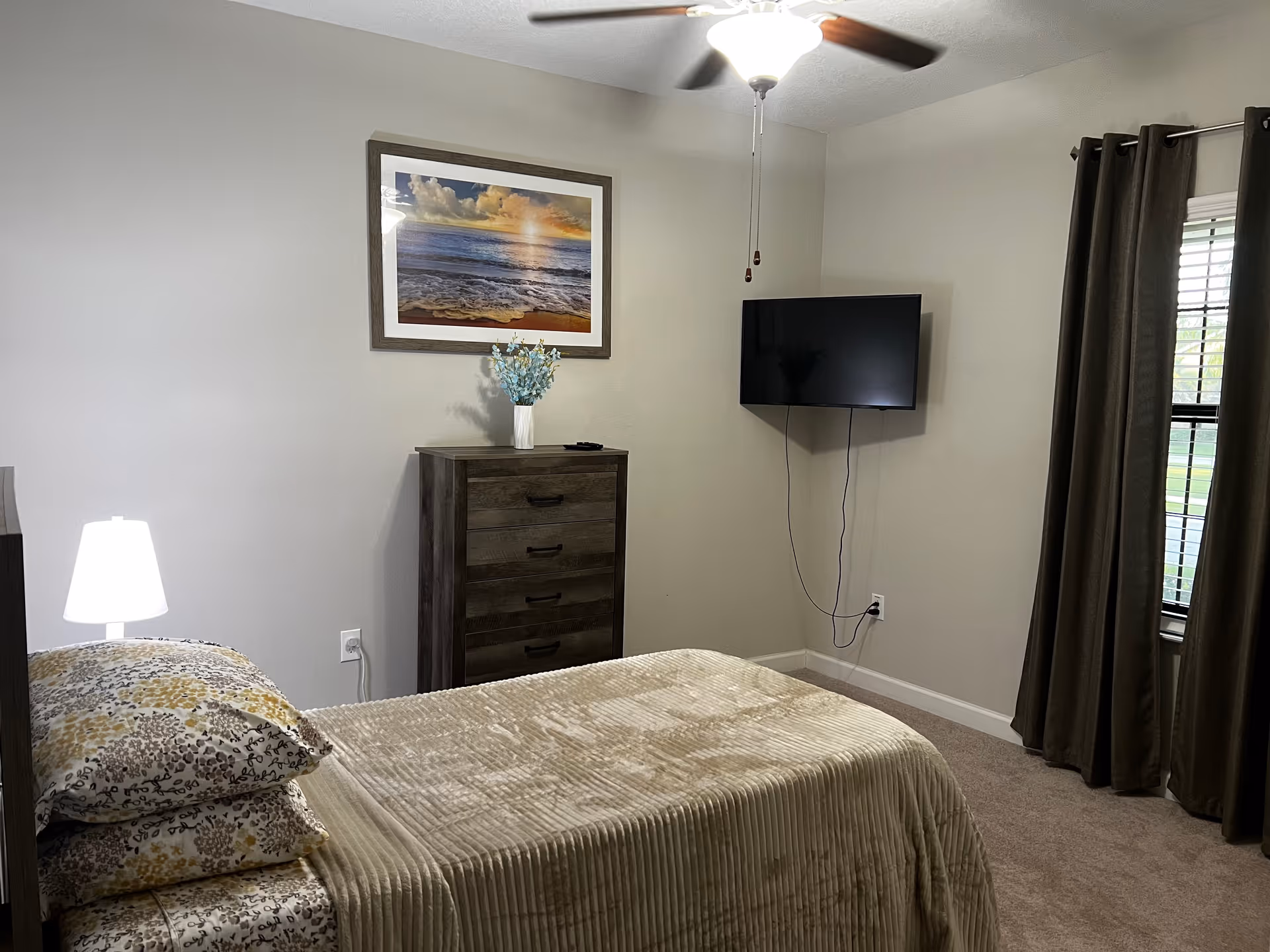 A bedroom in an assisted living facility featuring a single bed with patterned pillows and a beige blanket. There is a wooden dresser with a vase of flowers on top, a wall-mounted flat-screen TV, a ceiling fan with a light, and a window with dark curtains.
