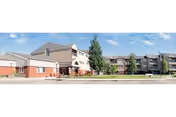 Exterior front view of a multi-story senior living facility with a driveway, landscaped lawn and trees under a blue sky.