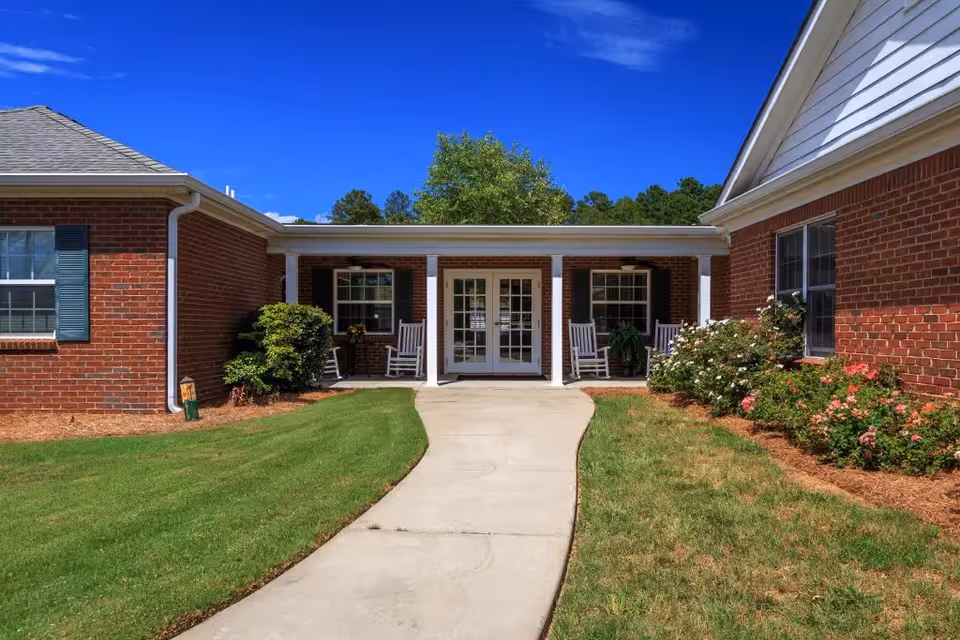 Brick assisted living building entrance with a curved walkway, white columns, rocking chairs on a covered porch, and landscaping under a blue sky.