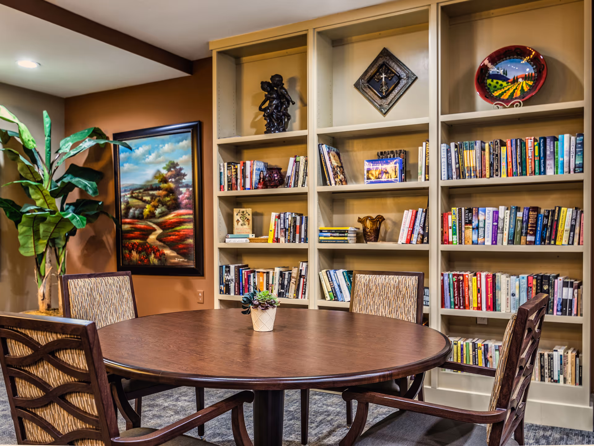 Round wooden table and chairs in a cozy reading area with built-in bookshelves, artwork, and a potted plant.