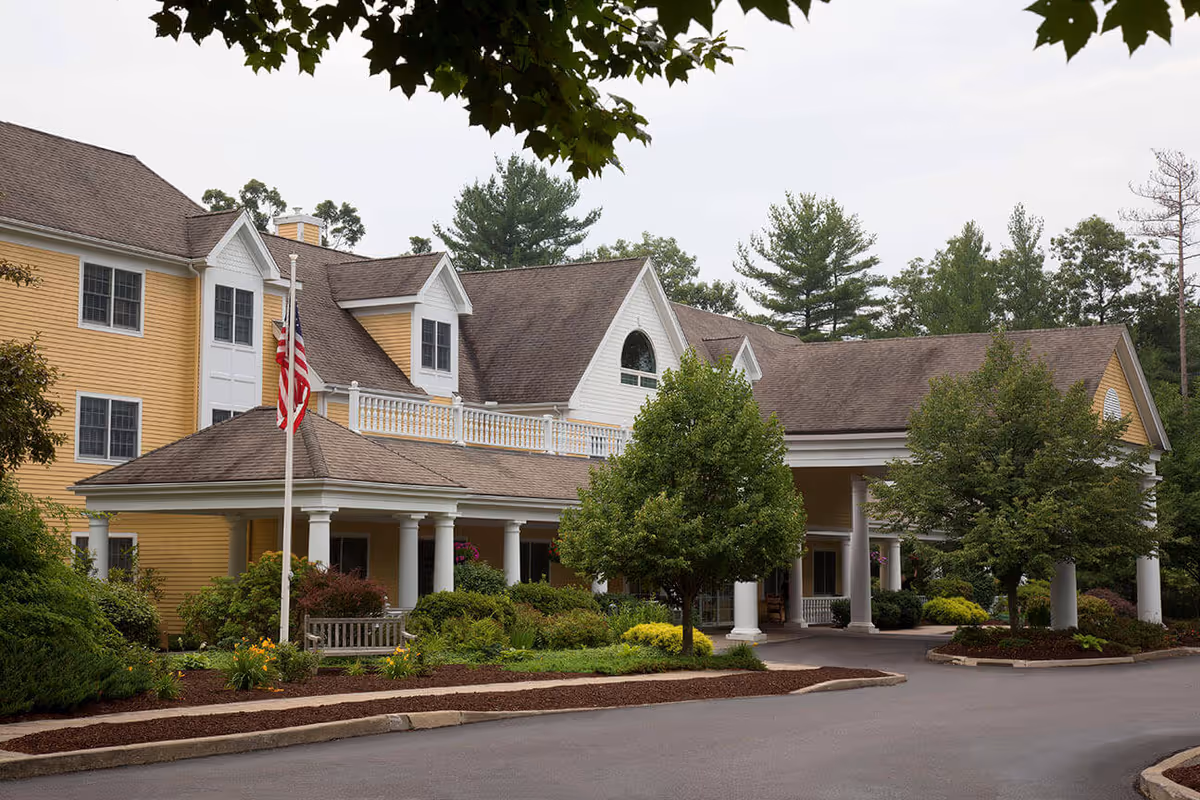 Front exterior of a yellow senior living building with a covered entrance, flagpole, and landscaped grounds.