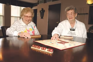 Two elderly women sitting at a table playing a game of Scrabble in a well-lit room with windows and dark wooden furniture.