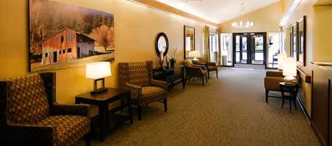 A warmly lit senior living lobby with armchairs, side tables, lamps, and framed wall art.