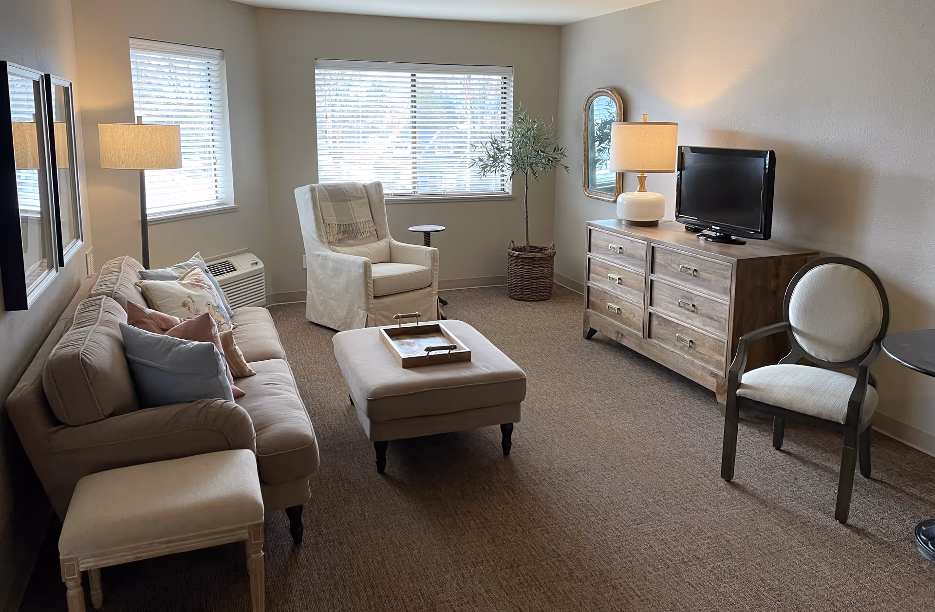 A bright living room with a sofa, armchair, ottoman, wooden dresser with TV, and lamps.