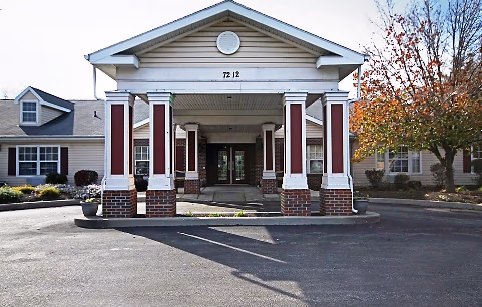 Front entrance of a senior living facility with a covered driveway supported by white columns with red panels and brick bases. The building has beige siding, multiple windows with dark shutters, and a tree with autumn-colored leaves on the right side.