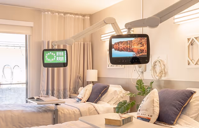 A bright shared bedroom with two beds, adjustable overbed tables, wall-mounted TVs, and bedside decor.