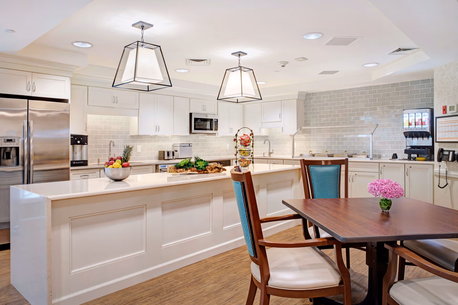 A bright, modern kitchen area in a senior living facility featuring white cabinetry, a large island with a bowl of fruit and a tiered snack basket, stainless steel refrigerator, microwave, toaster, and a beverage dispenser. There is a wooden dining table with chairs, one of which has a blue upholstered backrest, and a small vase with pink flowers on the table. The floor is wooden, and the walls have light gray subway tile backsplash.
