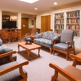 A cozy living room area in a senior living facility with multiple upholstered chairs and sofas arranged around a wooden coffee table. There is a bookshelf filled with books against the wall and a wooden dining table with chairs in the background. The room is well-lit with recessed ceiling lights.