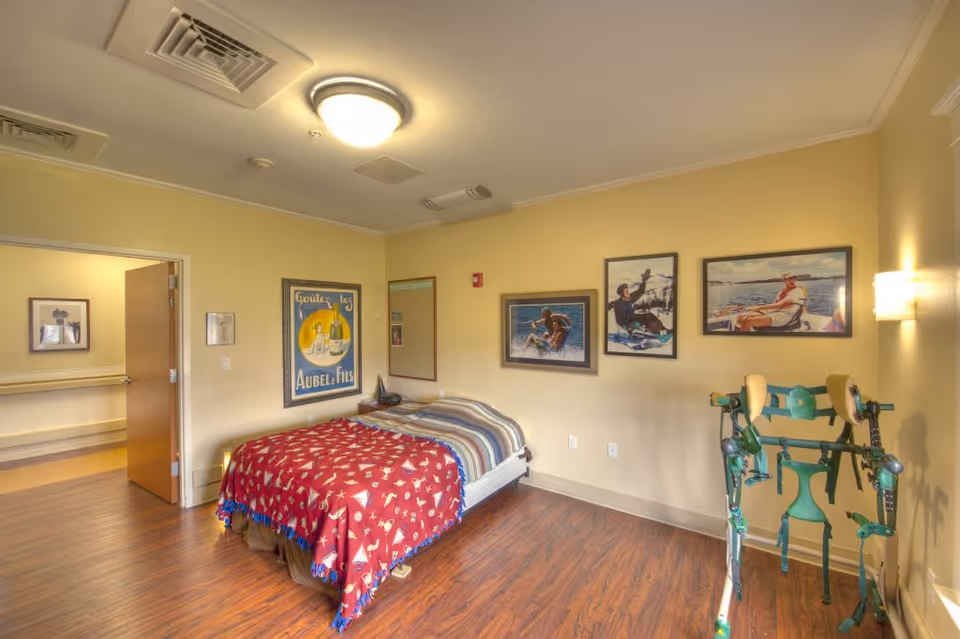 A bedroom in a senior living facility with a bed covered by a red blanket with white patterns and a striped sheet. The room has wooden flooring, beige walls, and several framed pictures on the walls. There is a green rehabilitation walker near the window on the right side. The door to the room is open, showing a hallway outside.